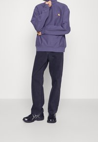 Henkilö, jolla on päällään violetti Carhartt-puolivetskari, mustat suorat farkut ja mustat Asics-tennarit, yksinkertaista valkoista taustaa vasten.