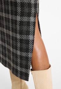 Grau-schwarz karierten Rock mit einem seitlichen Schlitz, aus einem Stoff mit weicher Textur. Ein Paar beige Overknee-Stiefel ist teilweise sichtbar.