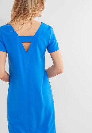 Femme portant une robe courte à manches courtes bleu vif avec un dos en V profond et une sangle horizontale, debout devant un fond clair uni.