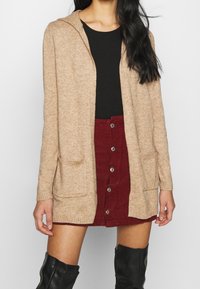 Béžový pletený cardigan s kapucí, předními kapsami a vroubkovanými manžetami. Nosit přes černý top a bordó manšestrovou sukni. Černé kozačky nad kolena.