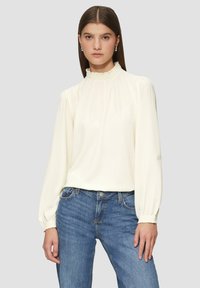 Blouse crème à col montant avec de longues manches bouffantes et un col volanté, associée à un jean en denim bleu. Tissu léger, texture douce.