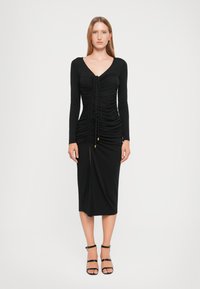 Black long-sleeve dress with ruching, V-neckline, front slit, and gold-button detail at the waist. Smooth fabric with a fitted silhouette.
