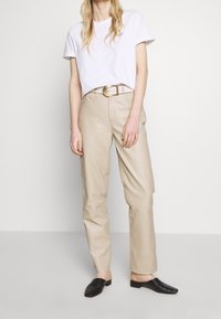 Beige läderbyxor, vit t-shirt och svarta texturerade klackar. Byxorna har en avslappnad passform och synlig sömnad, med ett guldskärp.