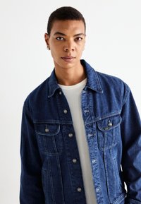 Persoon met kort haar die zilveren oorbellen van het type creolen draagt, een blauwe spijkerjacket over een witte gestructureerde blouse, recht vooruit kijkt tegen een witte achtergrond.