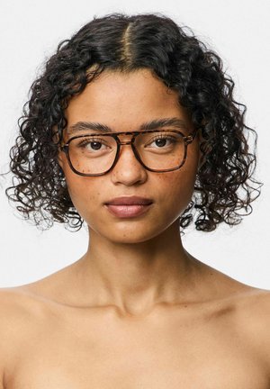 Ung person med kort, krøllet sort hår iført store briller med skildpaddeskalstel, set forfra med et neutralt udtryk mod en ensfarvet baggrund.