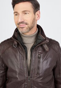 Bruine leren jas met een ritsluiting, hoge kraag en gequiltte schouderdetails. Heeft een borstzak met rits en een zachte binnenvoering.