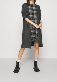 Šedý dlouhý kardigan přes černobíle kárované šaty, doplněný černými kotníkovými botami a bílými ponožkami. Jednoduché, texturované pletené.