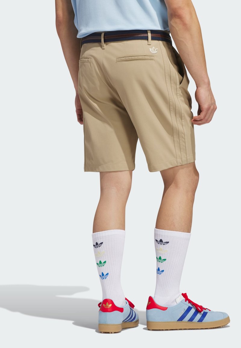 Homem a usar calções bege, meias brancas até ao tornozelo com logótipos multicoloridos da Adidas e sapatilhas Adidas azul claro com detalhes em vermelho.