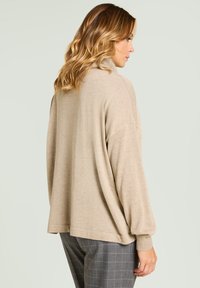 Beżowy, oversize'owy sweter z miękkiego dzianiny, z wysokim kołnierzem i prążkowanymi mankietami, zestawiony z szarymi spodniami w kratę. Gładka tekstura o luźnym kroju.