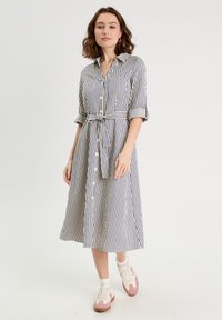 Robe chemise rayée en navy et crème, longueur genou, avec une taille cintrée, un devant boutonné et des manches retroussées. Associée à des baskets blanches et roses.
