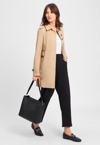 Beige trenchcoat met een ceintuur, knoopsluiting en zwarte broeken. Zwarte leren handtas met een vierkant ontwerp en schouderband.