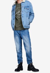 Chaqueta vaquera azul claro con cuello de borrego, sudadera verde y vaqueros azules desgastados, combinados con botas negras con cordones.