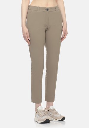 Pantaloni beige slim fit con chiusura a bottone indossati con sneakers beige e bianche su una persona in piedi su sfondo bianco.