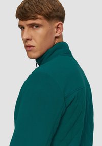 Teal fleece jack met een hoge kraag, geborduurde achterkant en ritssluiting. Gestructureerde stof en gestructureerde pasvorm. Racerback-stijl.