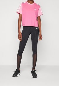 Rosa kortärmad t-shirt med vita ränder på ärmarna, tillsammans med svarta leggings med nätpaneler och Adidas-logotyp, bärs med svarta sneakers.