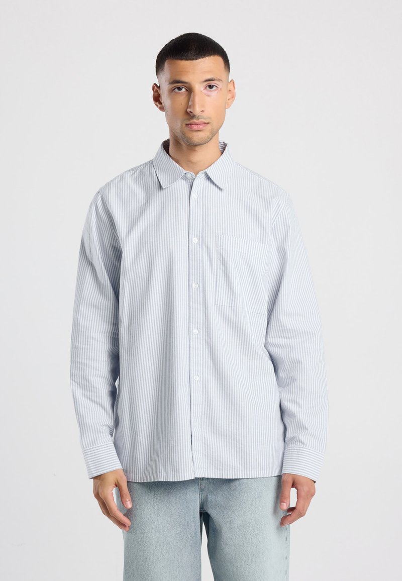 Camicia a maniche lunghe a righe blu chiaro con colletto button-down, tasca frontale e una texture sottile; abbinata a jeans di denim chiaro.
