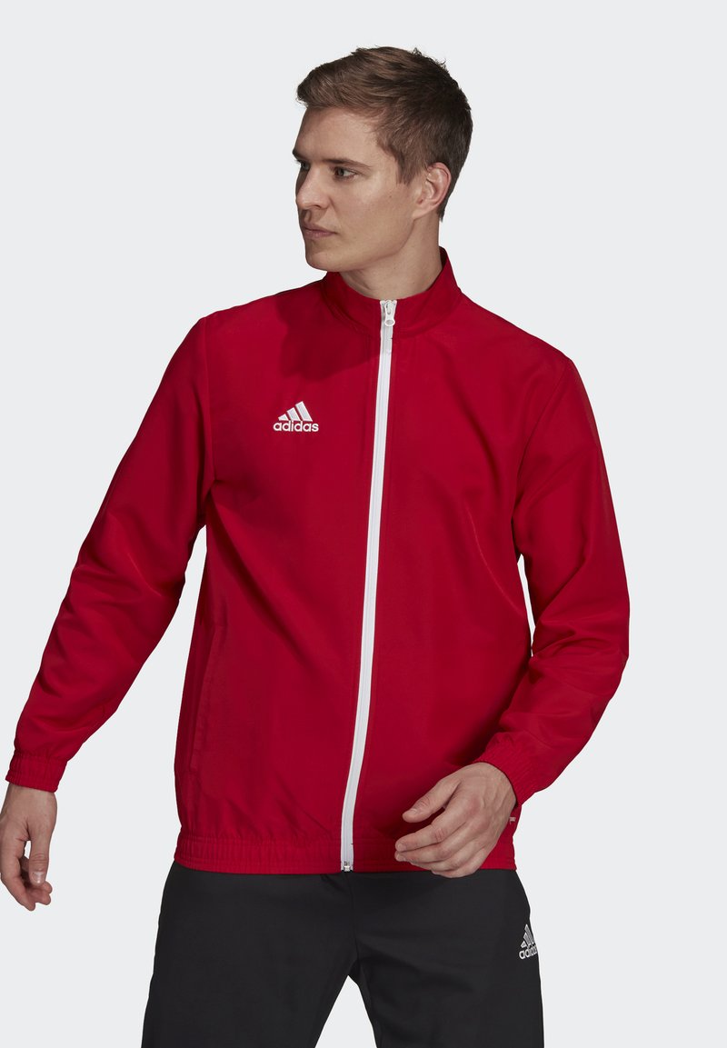 Rød træningsjakke med fuld lynlås, hvide detaljer, elastiske manchetter og sidelommer. Har et broderet logo på brystet.