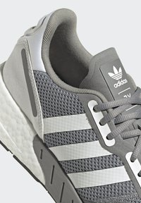 Šedá športová obuv s pleteným zvrškom, bielym trojprúžkom, polstrovaným golierom a textúrovanou gumovou podrážkou. Obsahuje označenie značky na jazyku.