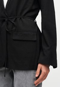 Μαύρο σακάκι με ζώνη γραβάτας και εμπρός τσέπη κεντημένη με το λογότυπο «KARL LAGERFELD JEANS», φορεμένο πάνω από γκρι τζιν, με το χέρι χαλαρό στο πλάι.