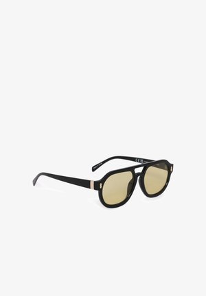 Lunettes de soleil à monture noire avec des verres teintés jaunes et un accent doré près des charnières, positionnées à un angle de trois quarts sur un fond blanc.