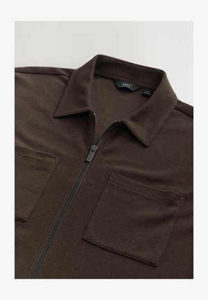 Veste zippée marron en tissu texturé avec un col, dotée de deux poches poitrine et d'une fermeture éclair noire. Étiquette visible à l'intérieur du col.