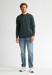 Donkergroene trui met een ronde hals en lange mouwen, gecombineerd met lichtblauwe jeans en witte sportschoenen. Casual pasvorm en eenvoudig ontwerp.