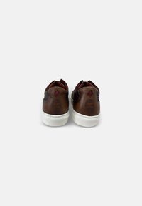 Zapatillas de cuero marrón con acentos texturizados, forro interior rojo y suela de goma blanca. Presenta un logo en forma de diamante y un diseño en relieve de una cara.