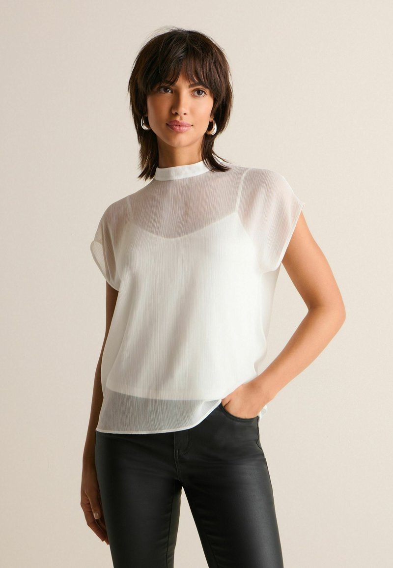 Femme avec des cheveux courts et foncés portant un chemisier blanc transparent à manches courtes sur un caraco blanc et un pantalon noir ajusté, posant avec une main dans sa poche.