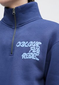 Zils džemperis ar rāvējslēdzēju un augstu apkakli, teksturēts audums, priekšpusē ar gaiši zilu izšūtu tekstu "COLOURFUL REBEL".
