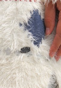 Bílá huňatá textilie s modrou pletenou nášivkou a šedým akcentem. Je viditelná černá značková etiketa. Ruka drží textilii.