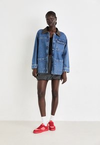 Giacca di denim con colletto marrone, quattro tasche anteriori, vestibilità oversized. Il modello indossa pantaloni corti grigi plissettati e sneakers rosse con calze bianche.
