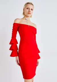Rød off-shoulder kjole med lange ermer som har lagdelte volanger. Tettsittende design, glatt tekstur, kne-lang passform. Utsmykket med en choker.