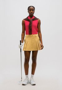 Kvinde iført lyserød ærmeløs top, beige plisseret nederdel, hvide sneakers, holder en golfkølle, med brun sweater draperet over skuldrene.