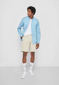 Camicia azzurra chiara a maniche lunghe sopra una maglietta bianca, abbinata a pantaloncini beige, sneakers bianche e calze con accenti neri. Trame lisce.