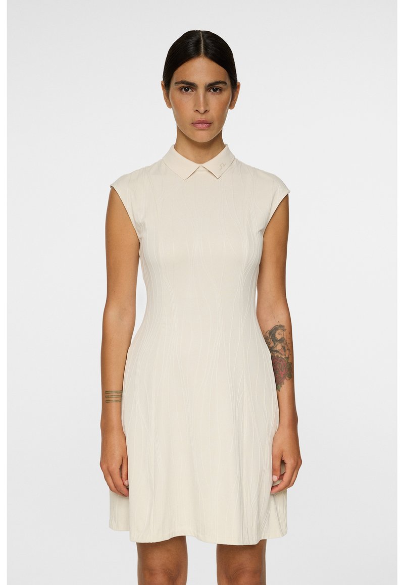 Nainen, jolla on tummat hiukset, pukeutuneena kermaiseen hihattomaan kauluspukuun, jossa on hienovaraisia pystysuoria kuvioita, seisoo vaaleaa taustaa vasten.