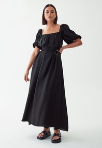 Black maxi dress with puff sleeves, square neckline, and a self-tie belt featuring a circular gold hardware detail. Textured fabric.