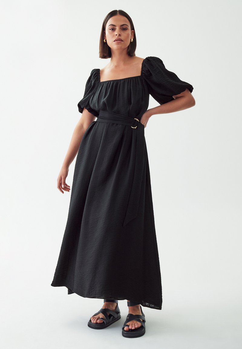 Black maxi dress with puff sleeves, square neckline, and a self-tie belt featuring a circular gold hardware detail. Textured fabric.