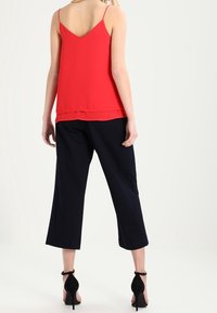 Donna che indossa un top rosso senza maniche, pantaloni neri a culotte e scarpe con tacco alto e cinturino alla caviglia nere, in piedi con le spalle alla macchina fotografica.