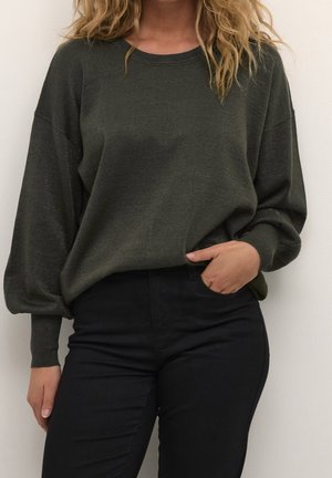 Femme portant un pull vert foncé à manches longues avec un pantalon noir, une main dans la poche, debout contre un mur blanc uni.