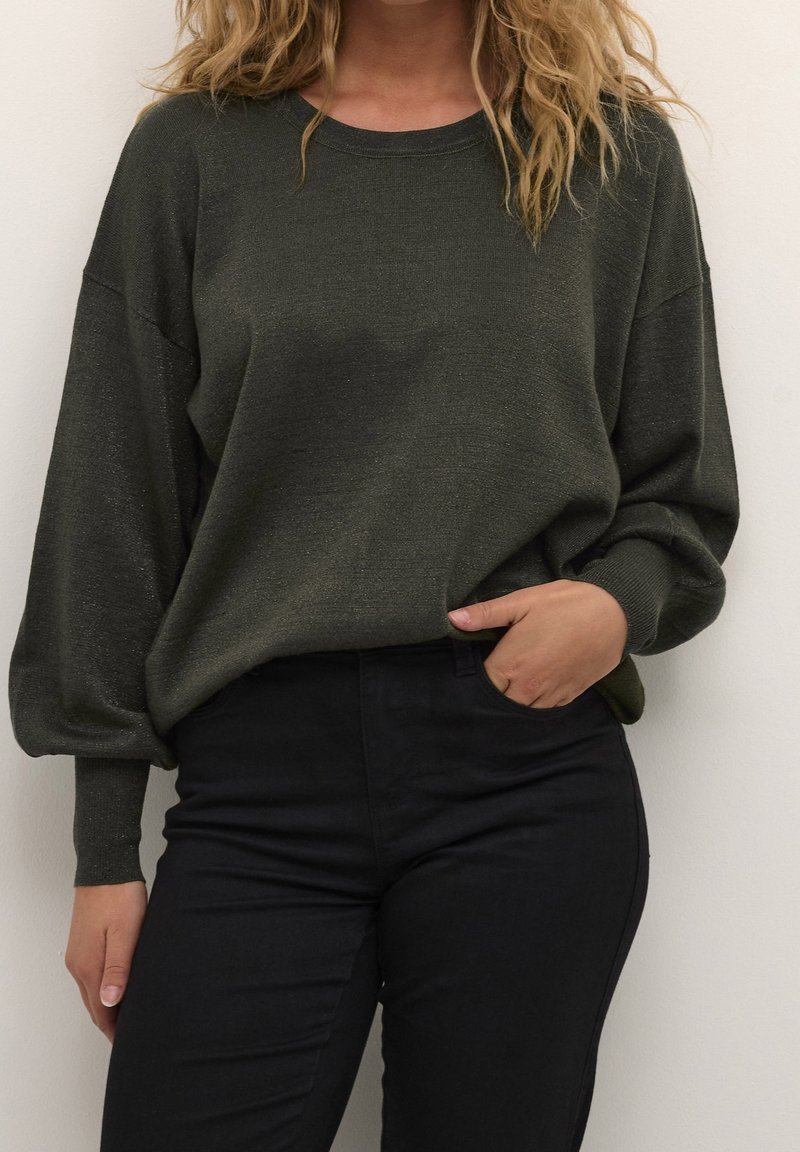 Femme portant un pull vert foncé à manches longues avec un pantalon noir, une main dans la poche, debout contre un mur blanc uni.