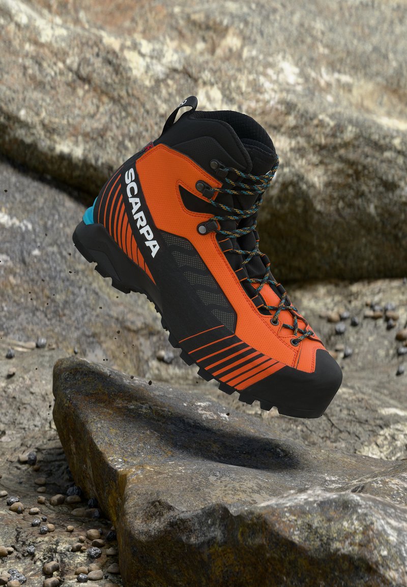 Πορτοκαλί και μαύρο μποτάκι πεζοπορίας με κορδόνια και ελαστική σόλα, με την ετικέτα Scarpa, τοποθετημένο πάνω σε βραχώδες έδαφος.