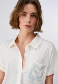 Polo blanc à manches courtes en tissu léger, doté d'une fermeture à boutons et d'une poche poitrine ornée de strass avec logo.