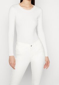 Mujer vistiendo un suéter ajustado de manga larga blanco de canalé y unos pantalones vaqueros blancos de cintura alta frente a un fondo simple.