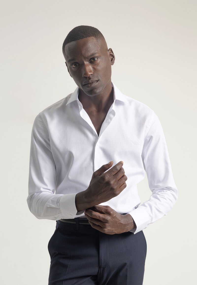 Camisa blanca de manga larga con cuello clásico, corte ajustado y puños con botones, combinada con pantalones oscuros de corte entallado. Textura de tela suave.