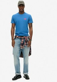 Blauwe katoenen t-shirt met een klein roze logo, lichtblauwe denim jeans, een geruit hemd om de taille geknoopt, grijze pet en zwarte schoenen.