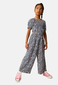 Blommig jumpsuit i marinblått med korta puffärmar, cinchad midja och vida ben. Har rosa blommönster. Matchas med rosa sneakers.