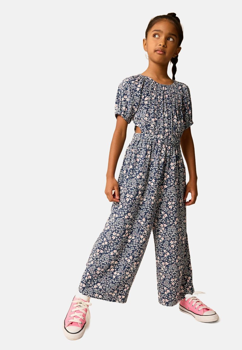 Blommig jumpsuit i marinblått med korta puffärmar, cinchad midja och vida ben. Har rosa blommönster. Matchas med rosa sneakers.
