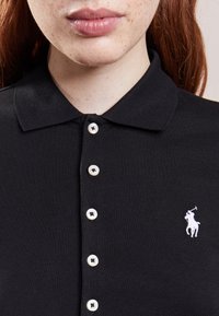Svart polo tröja med krage, som har vita knappar och en liten vit broderad logga av en h äst och ryttare på vänster bröst.