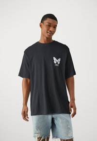 Black cotton t-shirt with short sleeves, featuring a white butterfly graphic on the left chest pocket area, paired with distressed denim shorts.