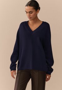 Dunkelblauer, übergroßer Pullover mit V-Ausschnitt, gerippten Akzenten und langen Ärmeln, kombiniert mit dunkelbraunen Lederhosen. Weiche Textur.